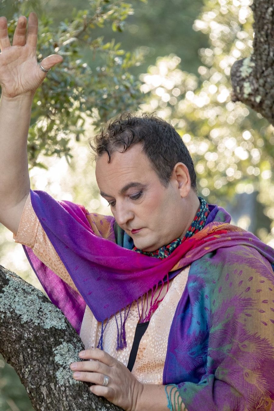 Un comédien en châle coloré, accoudé à une branche, en extérieur