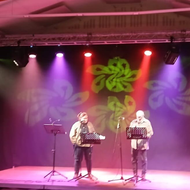 Deux comédiens au pupitre sur une scène montée en salle, projecteurs de couleur